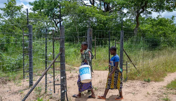 Two women walking along a sandy path beside a fenced rural area, carrying a water container.