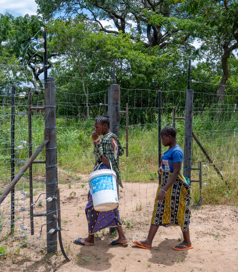 Two women walking along a sandy path beside a fenced rural area, carrying a water container.